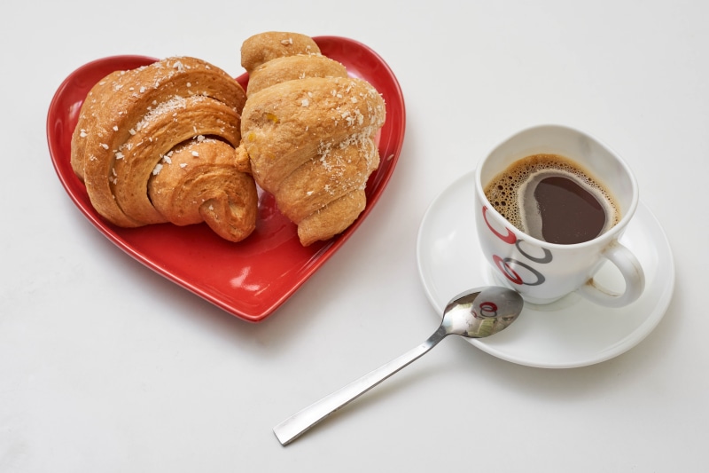 Two croissants on a heart-shaped plate with coffee and spoon