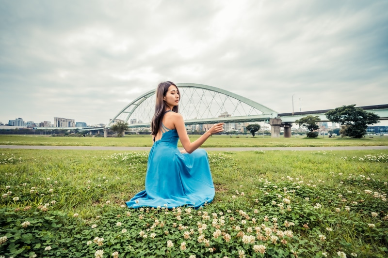 Asian woman in blue dress sitting on grass near bridge