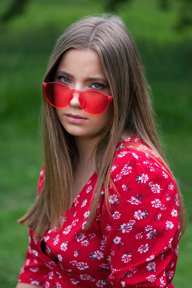 Girl with dark blonde hair wearing red heart-shaped glasses and a floral dress
