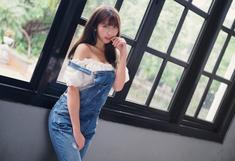 Asian woman in denim overalls posing by window