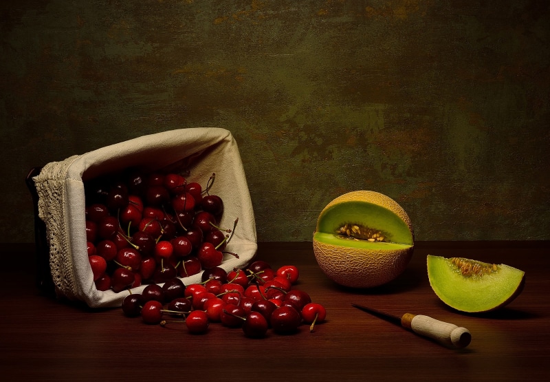 Cherries spilling from a basket with melon and knife