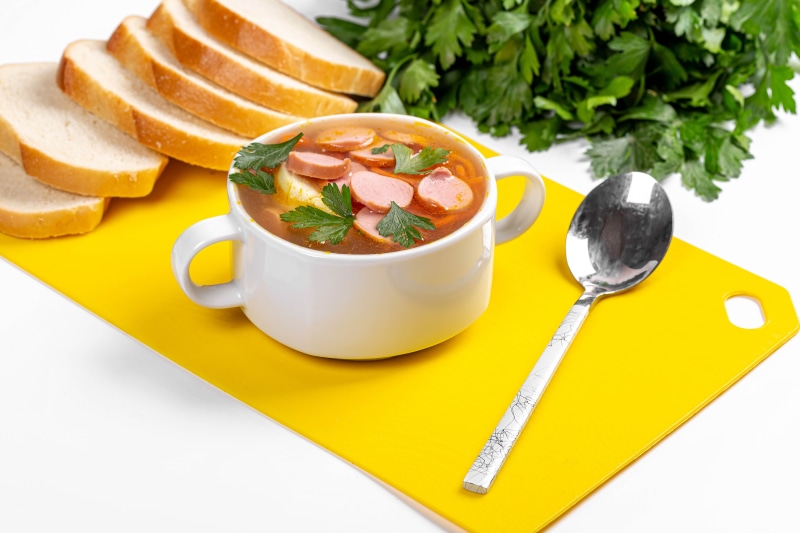 Soup with sausages, bread slices, parsley, and spoon on cutting board