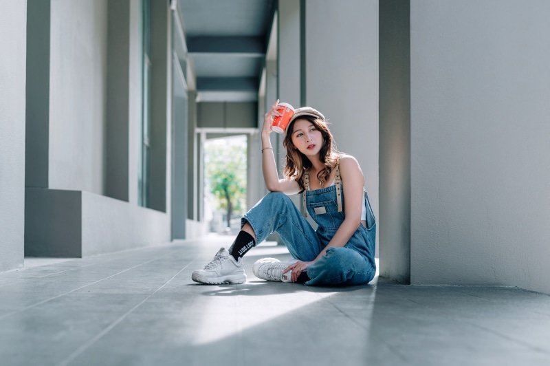 Asian woman wearing overalls and baseball cap sitting with drink