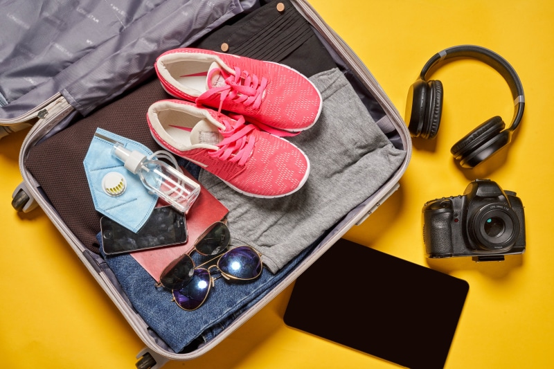Suitcase with travel essentials: mask, sanitizer, shoes, camera, headphones, tablet