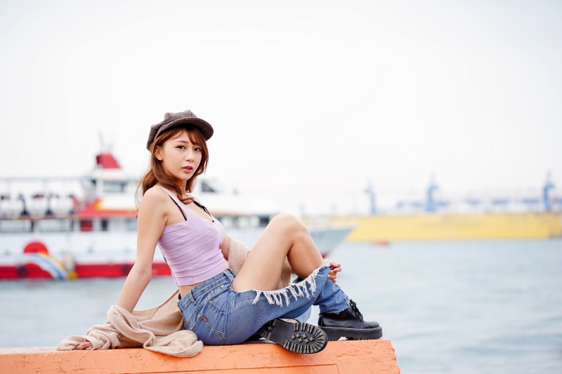 Asian woman wearing a baseball cap, singlet, and ripped jeans sitting by the water