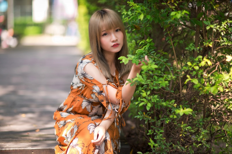 Asian woman in orange floral dress near green bush