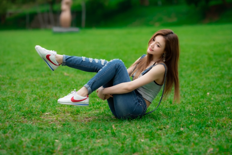 Asian woman in jeans and sneakers sitting on grass