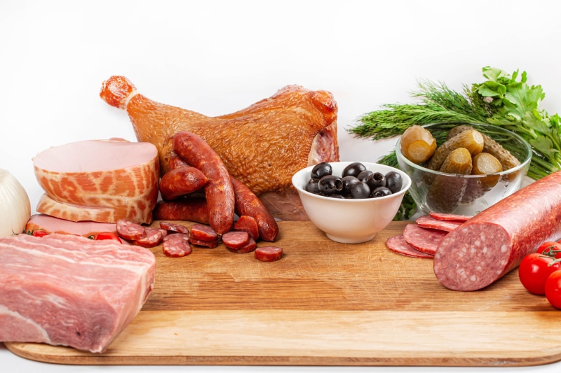 Meat products, sausages, ham, olives, pickles, and tomatoes on a cutting board