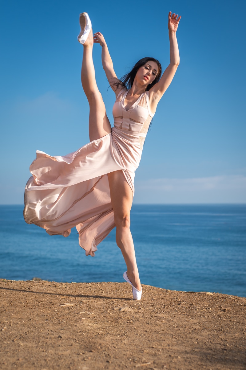 Ballerina in a flowing dress performs a high leg split by the ocean