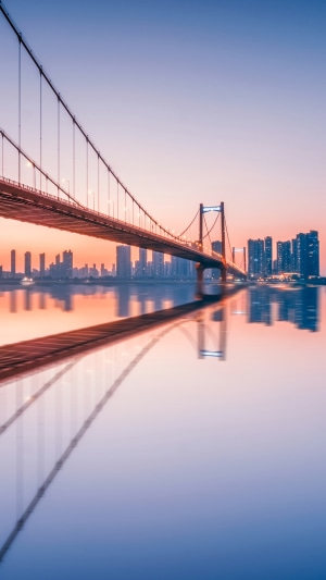 Suspension bridge over a river with city skyline at sunset