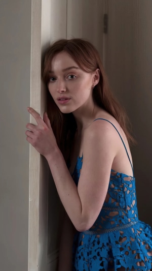 Phoebe Dynevor leans against a doorframe in a blue dress