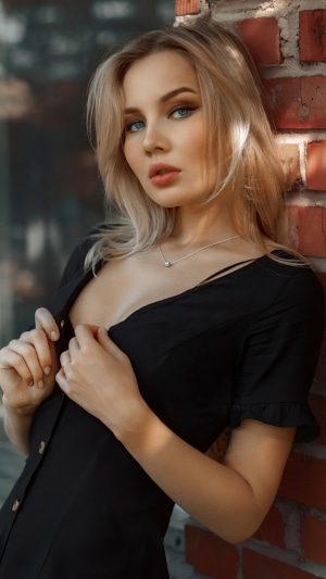 Blonde woman in black dress leaning against brick wall