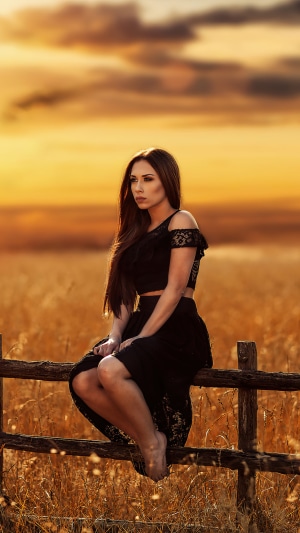 Woman in black dress sitting on a fence at sunset