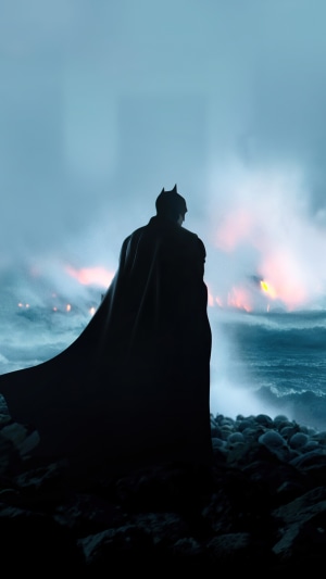 Batman standing on rocks with ocean and fire in background