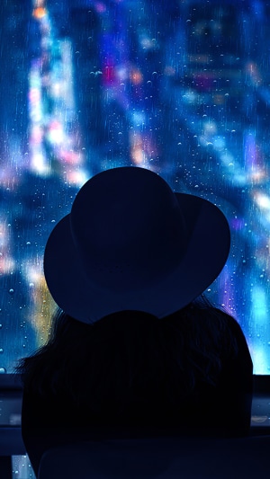 Silhouette of a person wearing a hat looking out a rainy window at city lights
