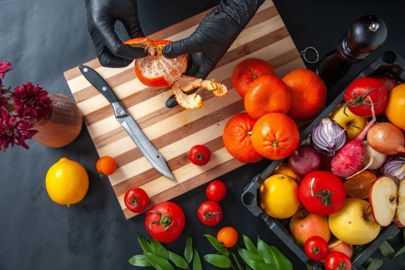 Hands peeling a tangerine on a cutting board with fruits and vegetables