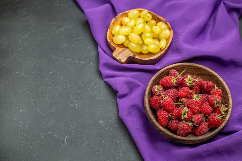 Raspberries and grapes in bowls on purple cloth