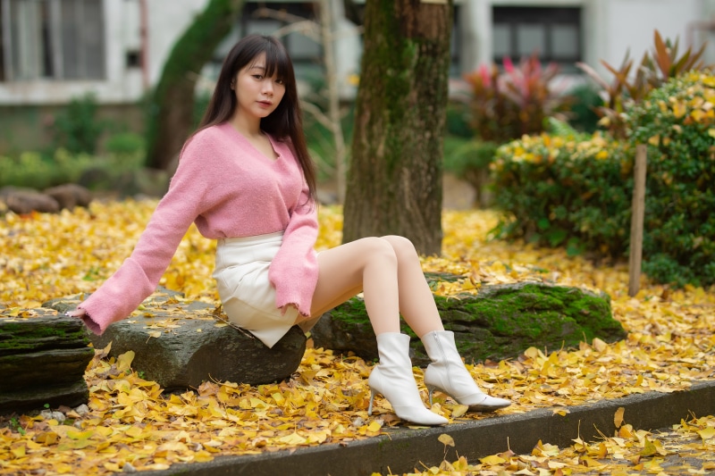 Asian woman in pink sweater and white skirt sitting in autumn foliage