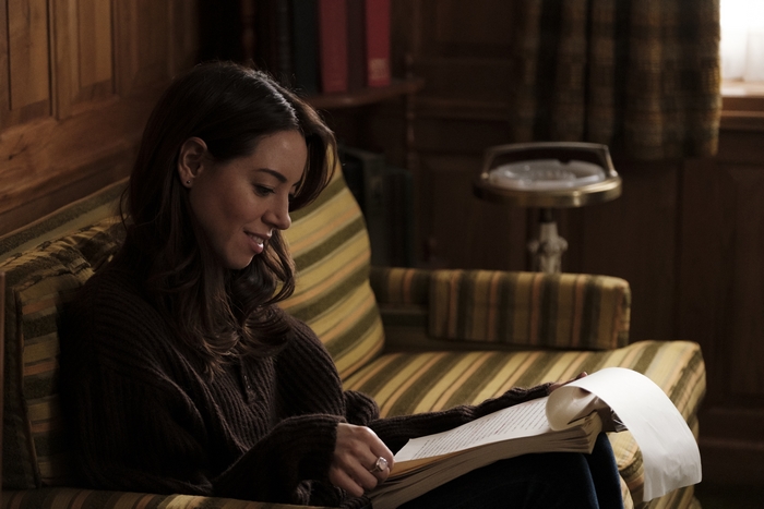 Aubrey Plaza smiling while reading a book on a couch