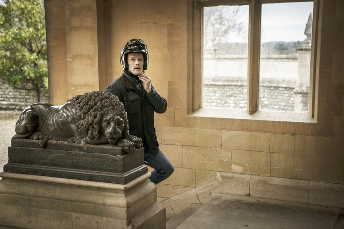 Sam Heughan adjusting helmet next to lion statue