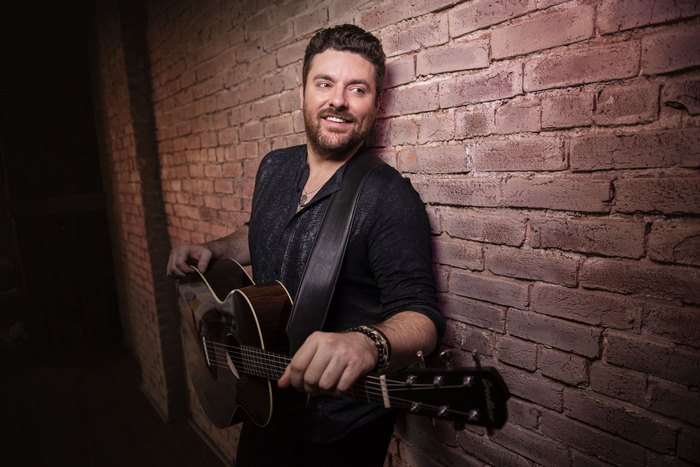Chris Young smiling with acoustic guitar against brick wall
