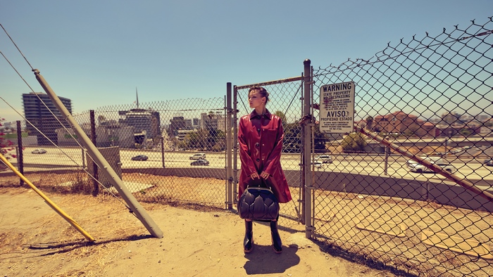 Olivia Holt in red coat by urban highway fence