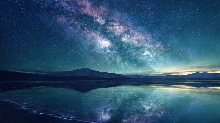 Milky Way galaxy reflected over a calm body of water with mountains in the background at night