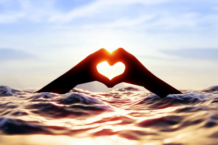 Hands forming a heart shape over the ocean at sunset