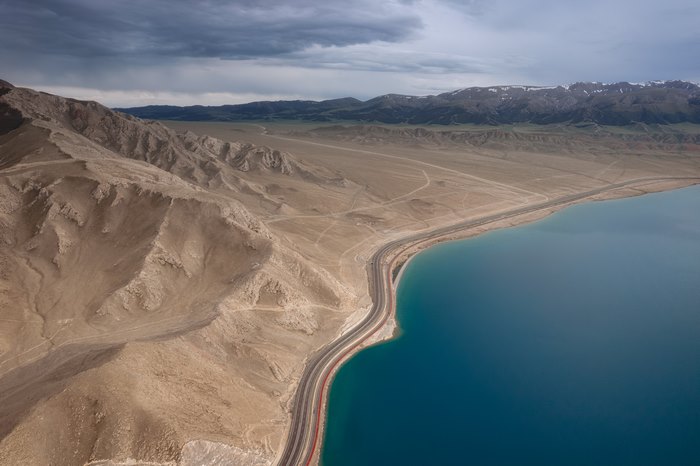 Aerial view of a highway along a vast blue lake and barren mountains