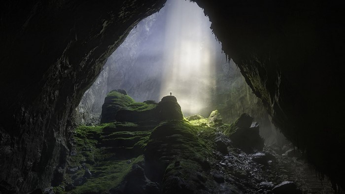 Person standing on mossy rocks in a cave with sun rays