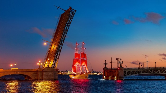 Sailing ship with red sails passes under a raised bridge at dusk in St. Petersburg