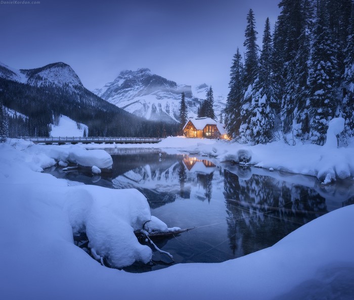 Winter cabin by frozen lake with snow-covered mountains