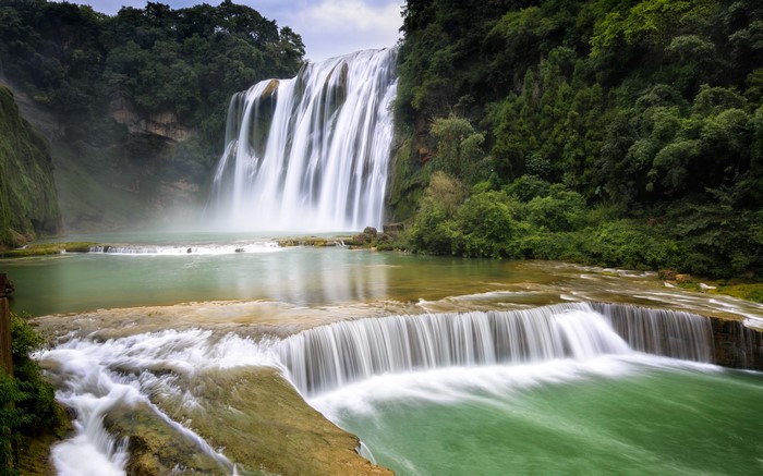 A wide waterfall cascades into a turquoise pool surrounded by lush green trees.