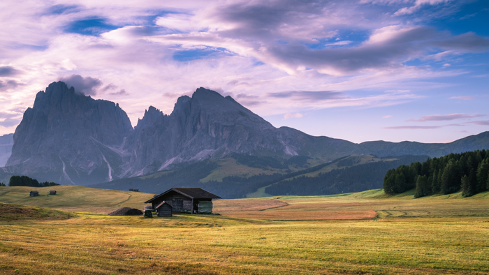 Wooden cabin in a grassy field with mountains and clouds