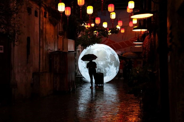 Person with umbrella and luggage in front of moon art installation at night
