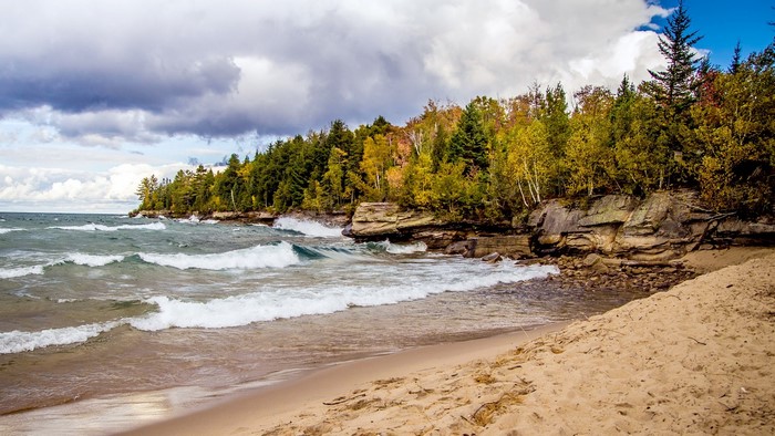 Waves crashing on a sandy beach with rocky cliffs and autumn trees