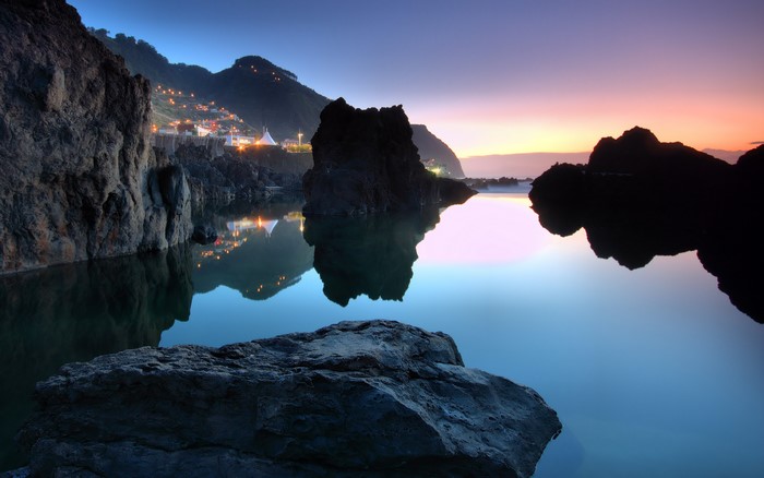 Rocky coastline with town lights reflecting in water at dusk