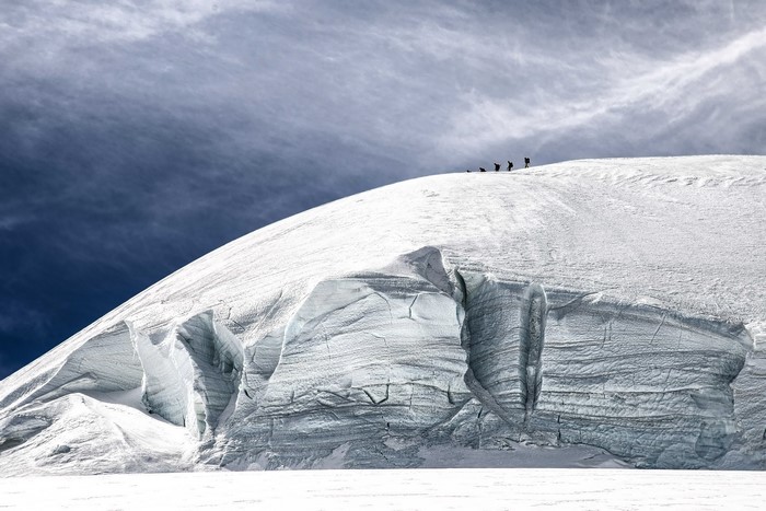 Climbers ascend a snow-covered mountain peak under a cloudy sky.