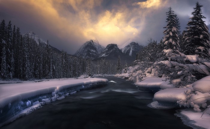 Winter river flowing through snow-covered forest and mountains under cloudy sky