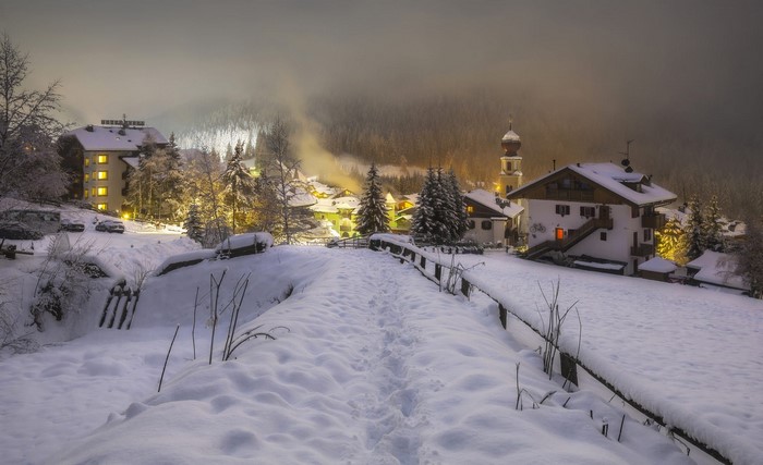 Snowy Italian village at dusk with illuminated buildings and church tower
