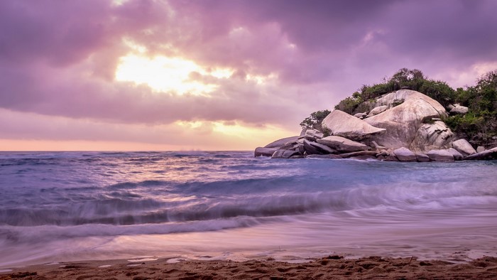 Long exposure of waves crashing on a beach with rocks and trees at sunset