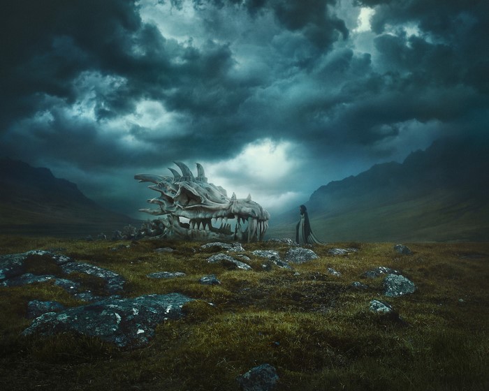 A person in a cloak stands by a dragon skull in a rocky field