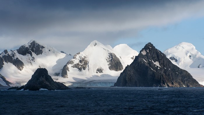 Snowy mountains and dark rocks in the ocean under clouds