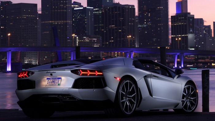 Silver Lamborghini Aventador parked by a city skyline at dusk