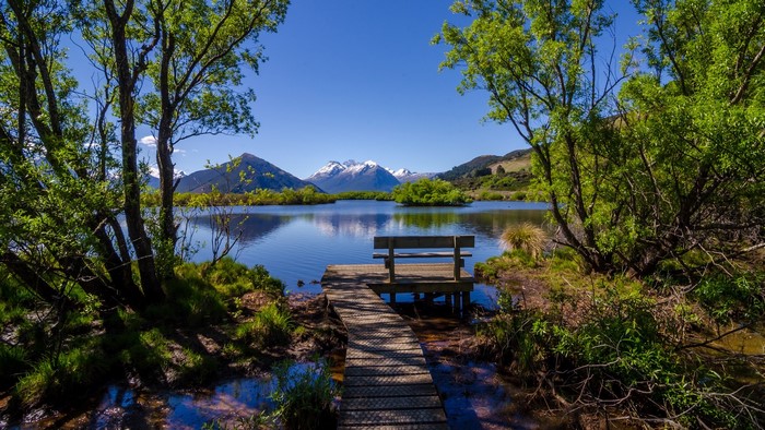 Wooden walkway and bench by a lake with snowy mountains
