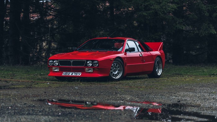 Red Lancia 037 rally car parked near trees with reflection