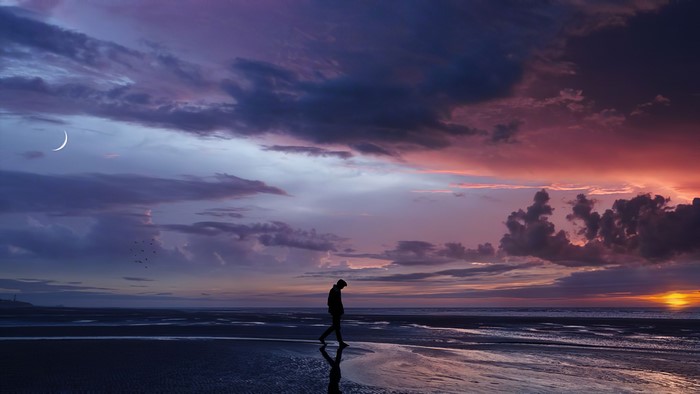 Person walking on a beach at sunset with crescent moon