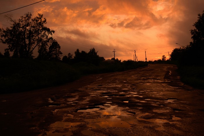 Potholed road reflecting sunset sky with trees and power lines