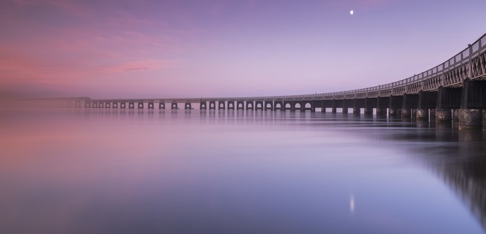 Long bridge over calm water at sunrise with moon