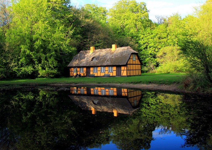 Thatched roof house reflected in a calm lake surrounded by trees